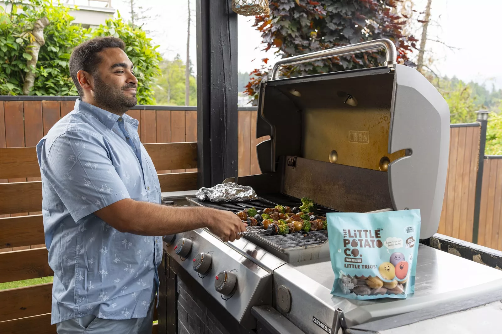 A man standing in front of a grill cooking Little Potatoes on a skewer, smiling at his family.